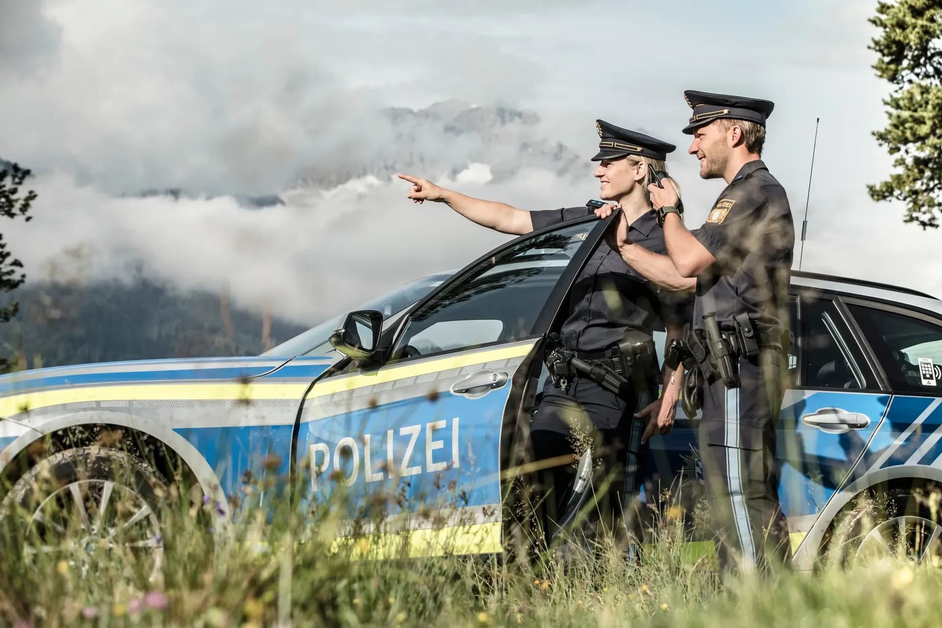 Zwei Polizeibeamte am Streifenwagen zeigen auf ein Bergpanorama.