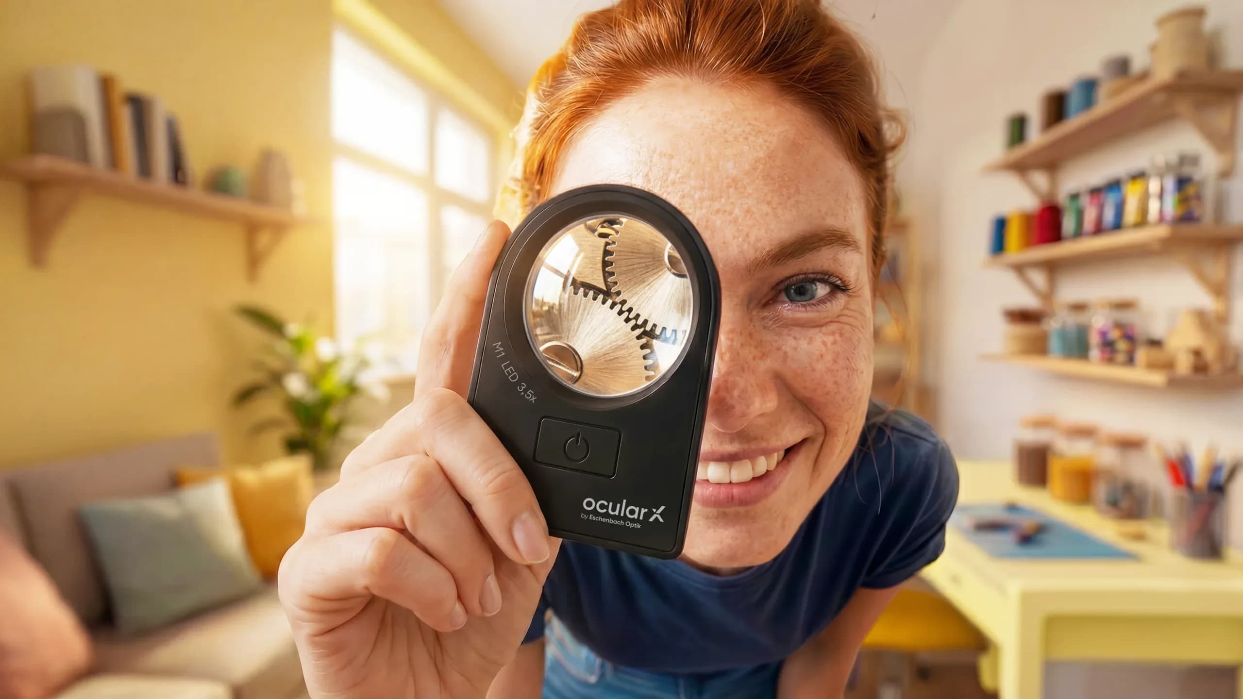 Eine Frau hält eine OCULAR-X-Lupe vor ihr Auge und lächelt, hinter ihr ein Hobbyraum mit Regalen