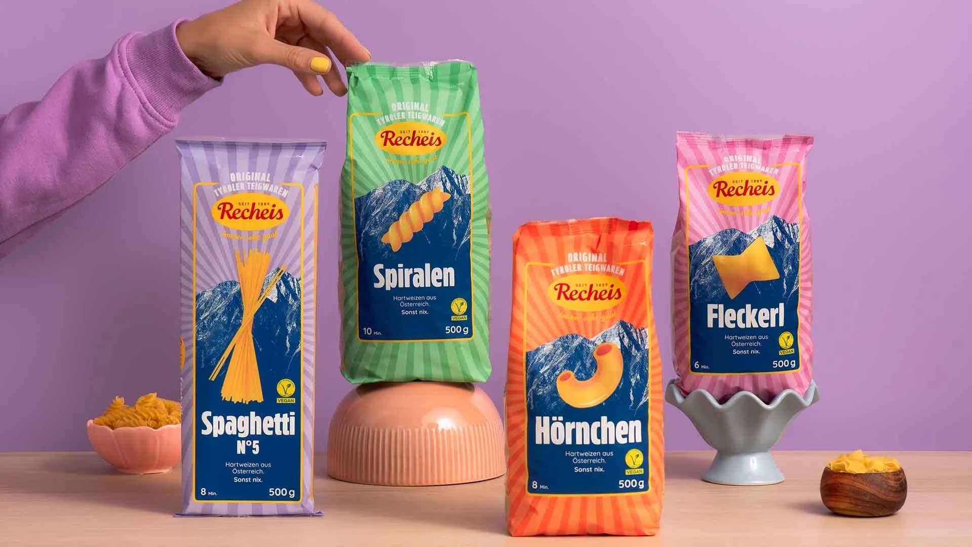 Four Recheis packs against a purple wall: Spaghetti N°5, Spiralen, Hörnchen and Fleckerl, with a hand reaching for the Spiralen